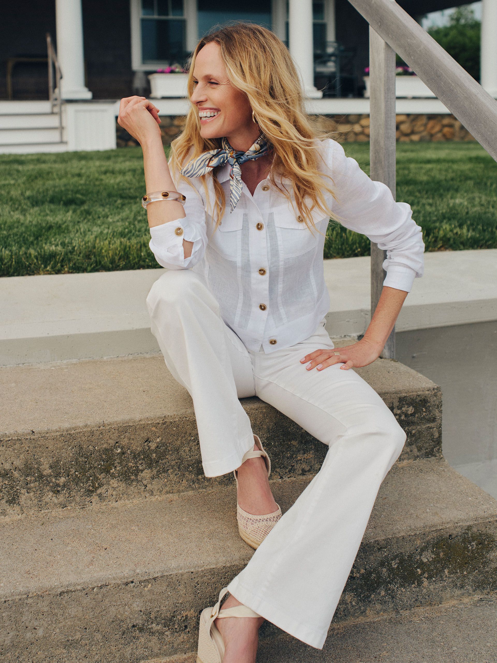 A smiling woman with blonde hair, wearing a white linen shirt, white pants, and a neck scarf, sits on concrete steps.