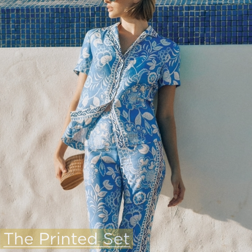 woman leaning against white stucco wall wearing blue paisley printed silky short sleeve button down with matching pants