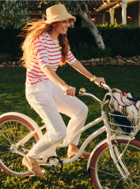 A smiling woman in a straw hat, red and white striped shirt, and white pants rides a white bicycle with a basket across sunlit grass.
