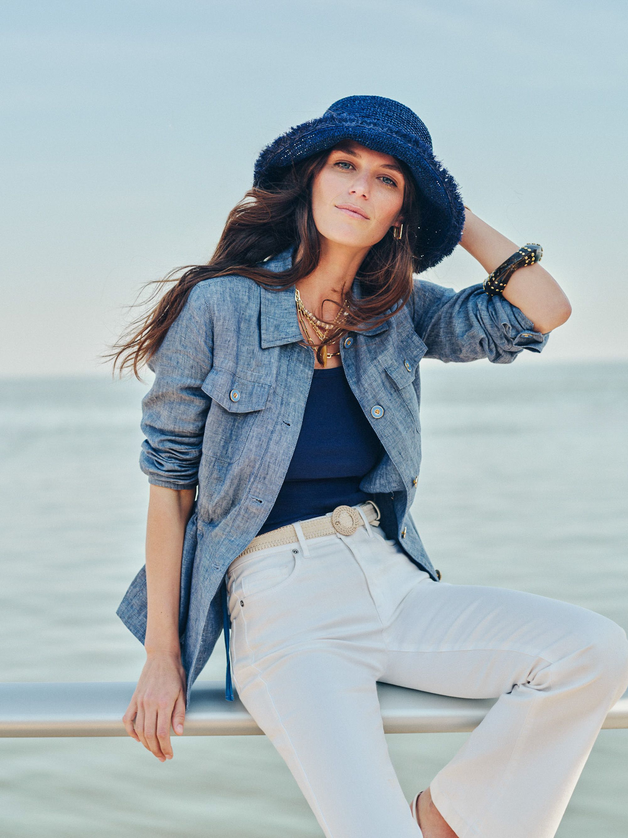 A woman wearing a blue textured hat, blue jacket, and white pants sits on a railing with the ocean behind her.