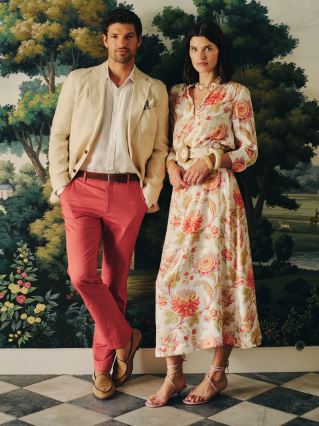 A man in a beige blazer and coral pants stands with a woman in a long cream floral dress, against a nature mural.