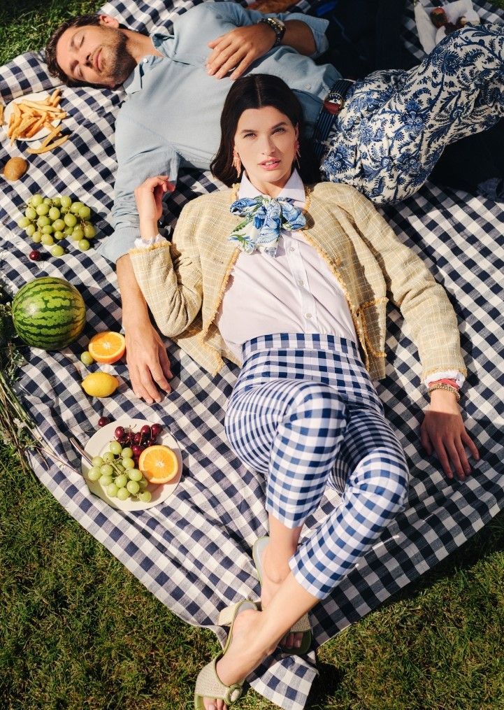 A woman in blue gingham pants looks at the camera, lying on a blue and white gingham picnic blanket next to a resting man, with fruit and fries.