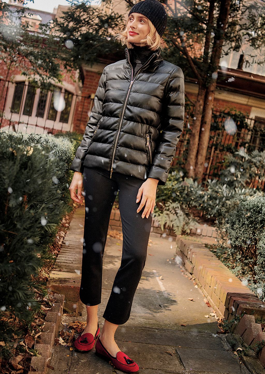 A woman wearing a black faux leather puffer jacket, black turtleneck, black pants, black beanie and red velvet loafers,=.