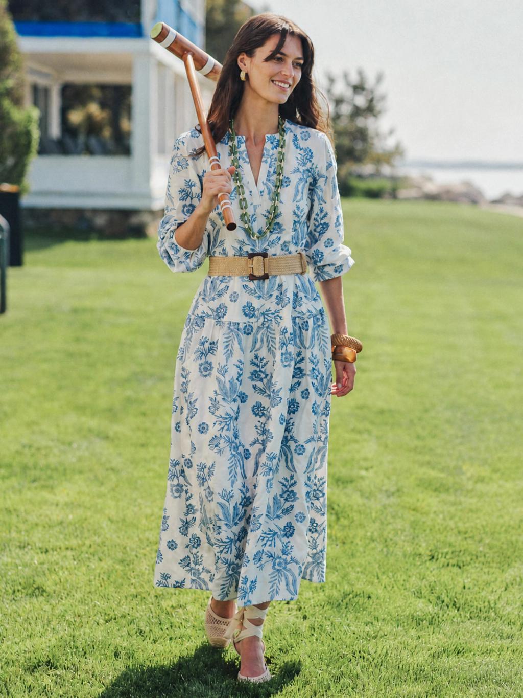 A smiling woman in a blue and white floral dress walks across a sunny lawn, carrying a croquet mallet over her shoulder.