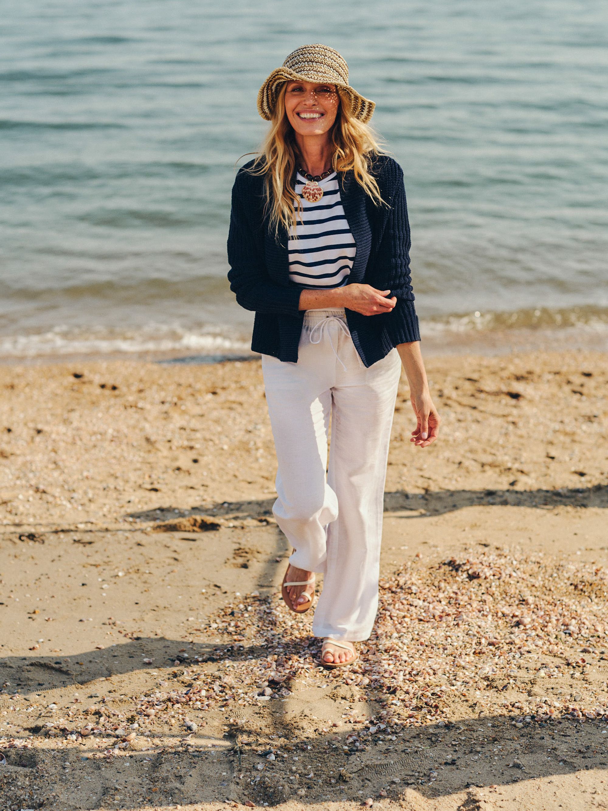 A smiling woman in a straw hat, striped top, and white pants walks on a sandy beach by the ocean.