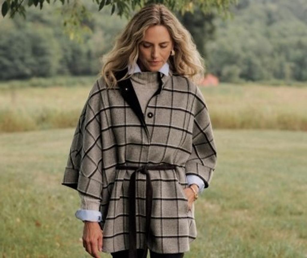 A woman with blonde hair wears a beige and black plaid belted poncho over a light shirt, standing in a grassy field.