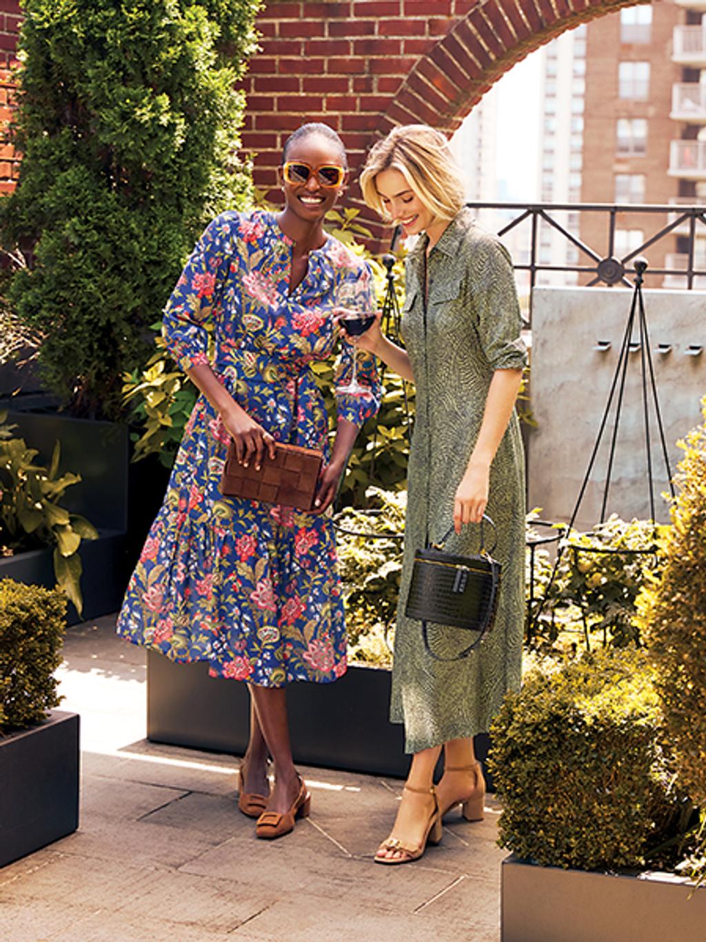 Two women wearing floral and patterned dresses and heels standing together, smiling while holding handbags.