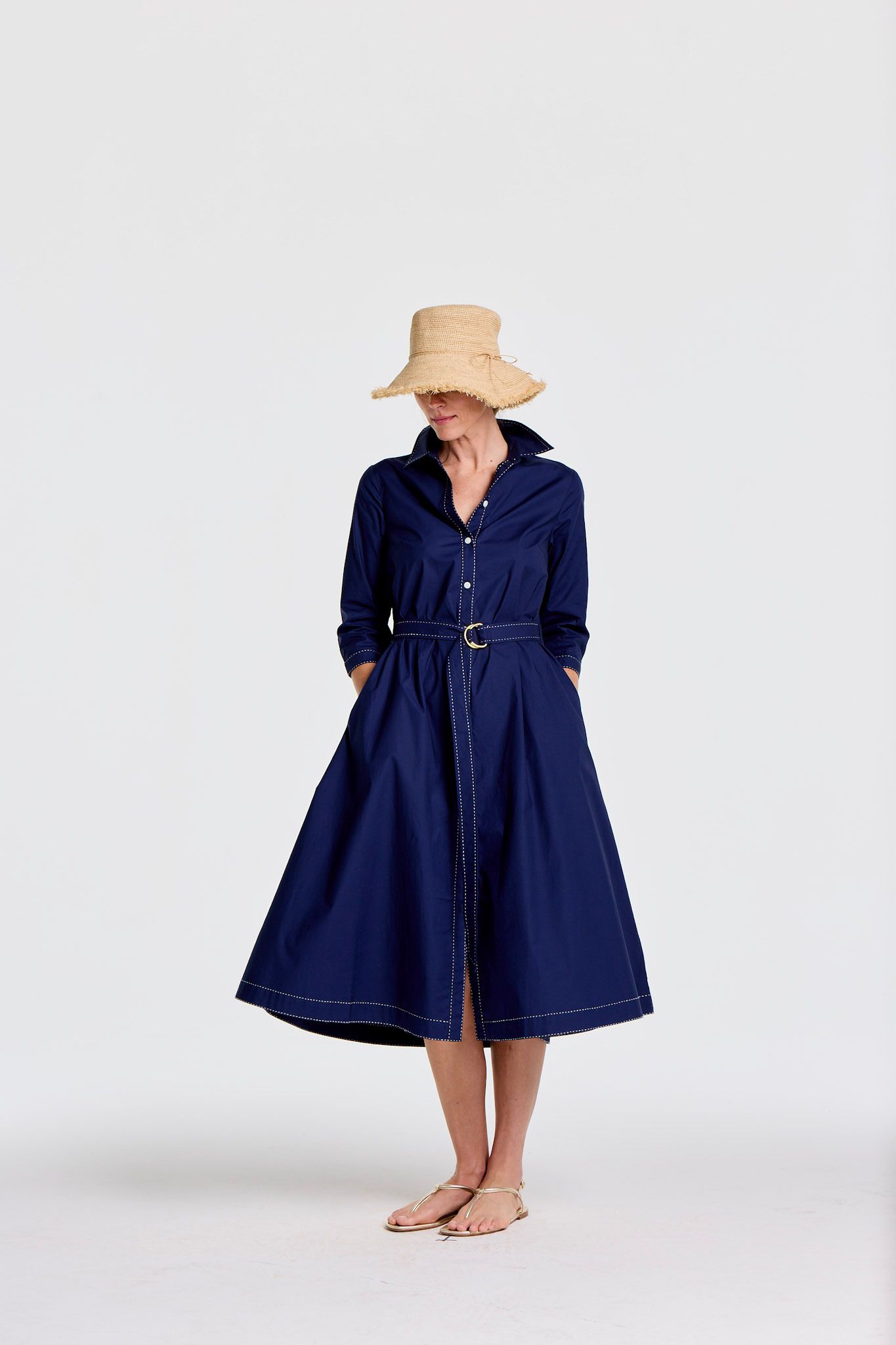 A woman wearing a straw hat, a navy blue shirtdress with white stitching and a belt, and gold sandals.