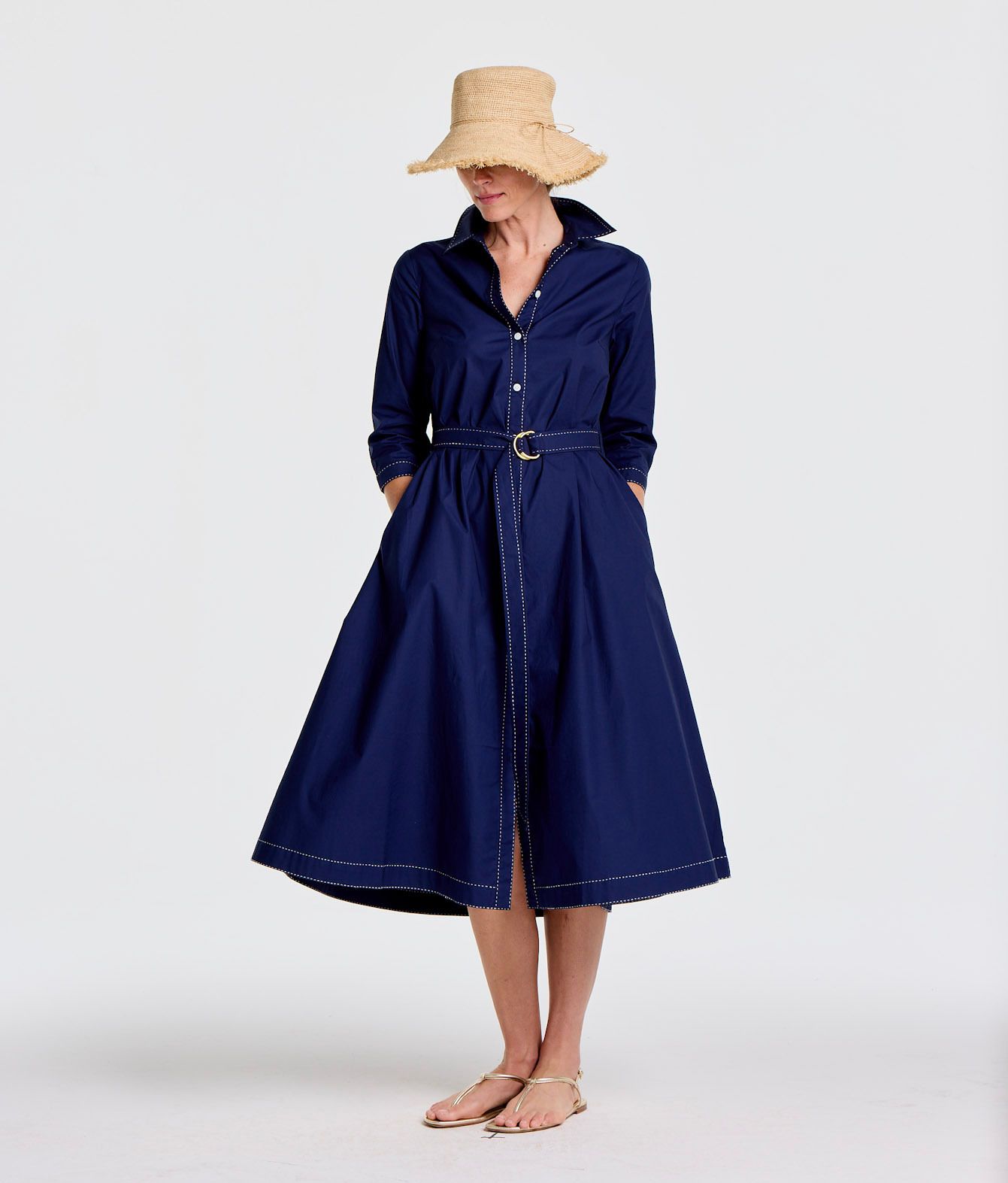 A woman wearing a straw hat, a navy blue shirtdress with white stitching and a belt, and gold sandals.