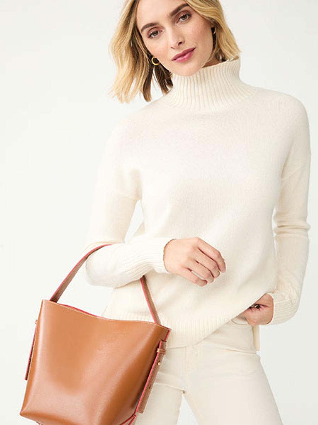 Model wearing white turtleneck and pants, carrying a brown bucket bag Model wearing white turtleneck and pants, carrying a brown bucket bag