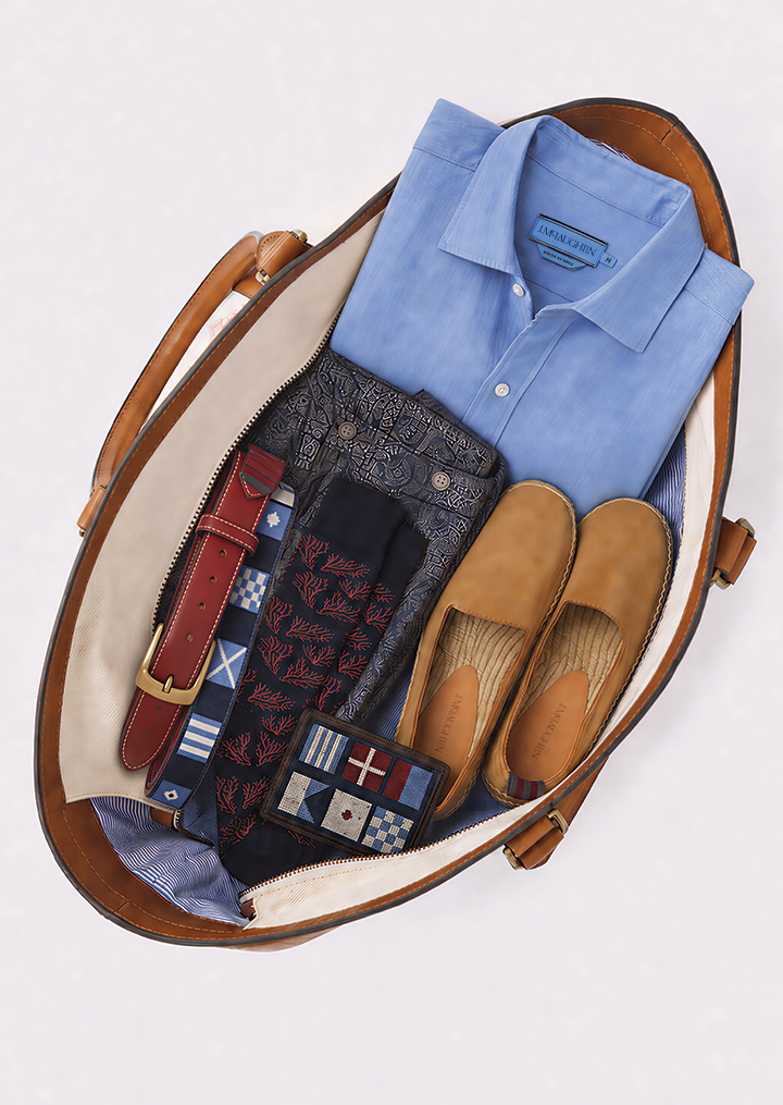 Brown leather tote bag containing men's summer clothing: a blue shirt, patterned shorts, red belt, socks, tan espadrilles, and a flag wallet.