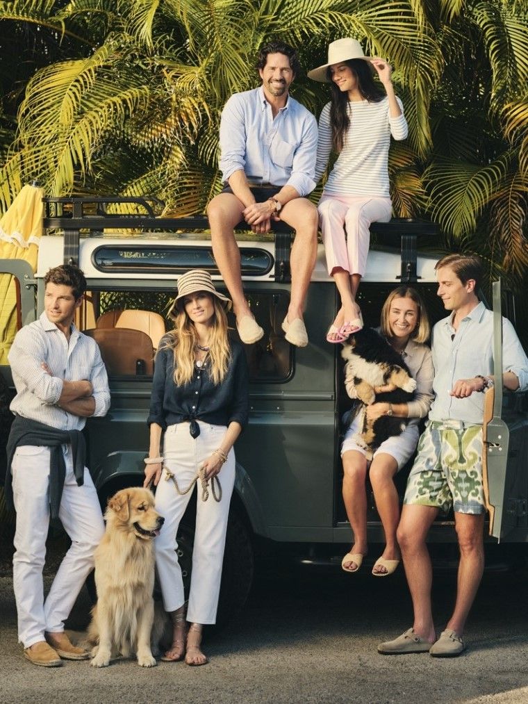Six people and two dogs pose around a vintage green SUV with palm trees in the background.