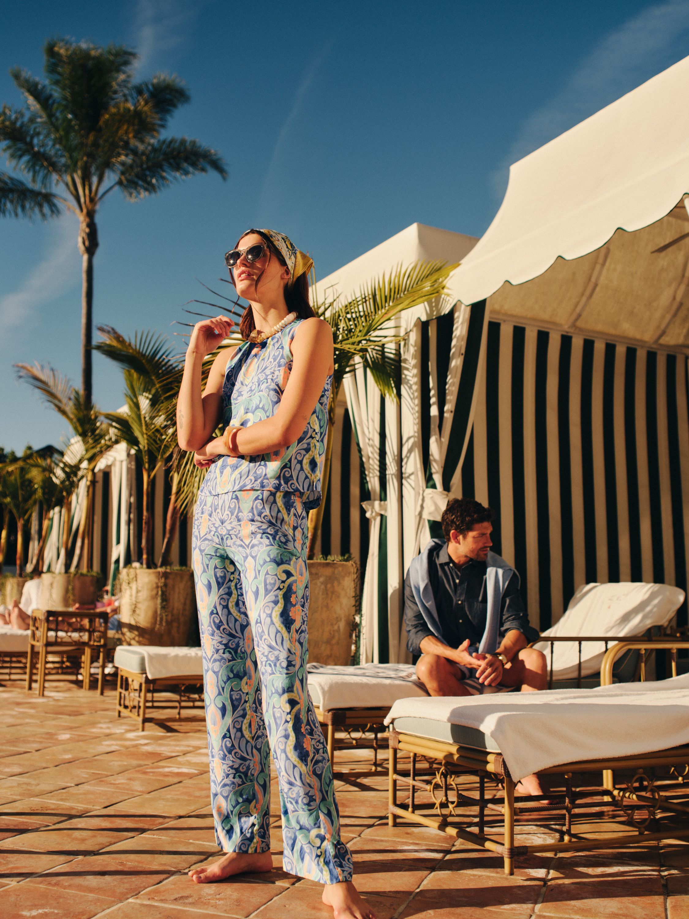 A woman in a vibrant patterned resort outfit, sunglasses, and headscarf stands on a sunny patio, with cabanas, palm trees, and a man on a lounger in the background.