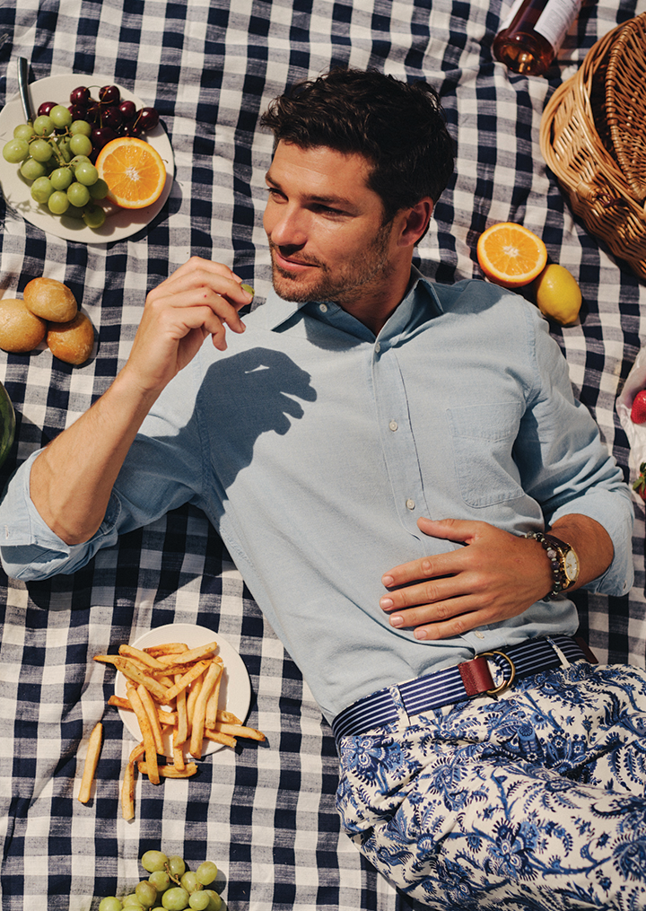 A man lies on a blue and white checkered picnic blanket, holding a grape and surrounded by various foods.