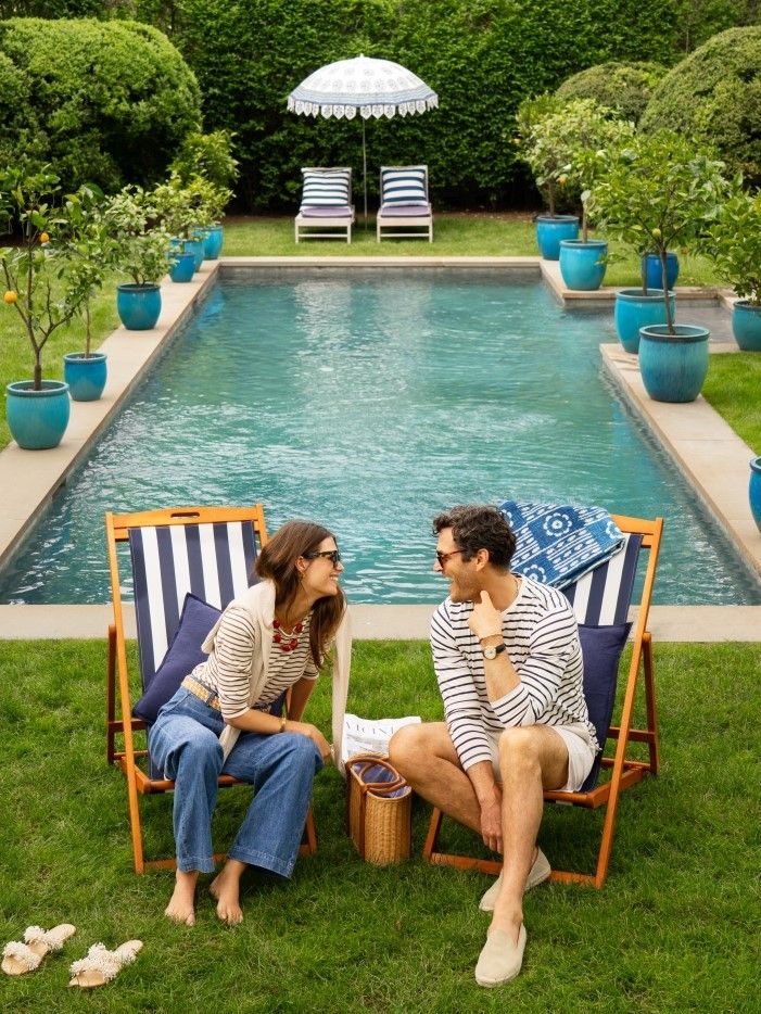 woman and man sitting outside in adirondak chairs by a pool, she is wearing jeans and brown and cream striped tee, he is wearing navy and cream striped tee and stone shorts with suede espadrilles