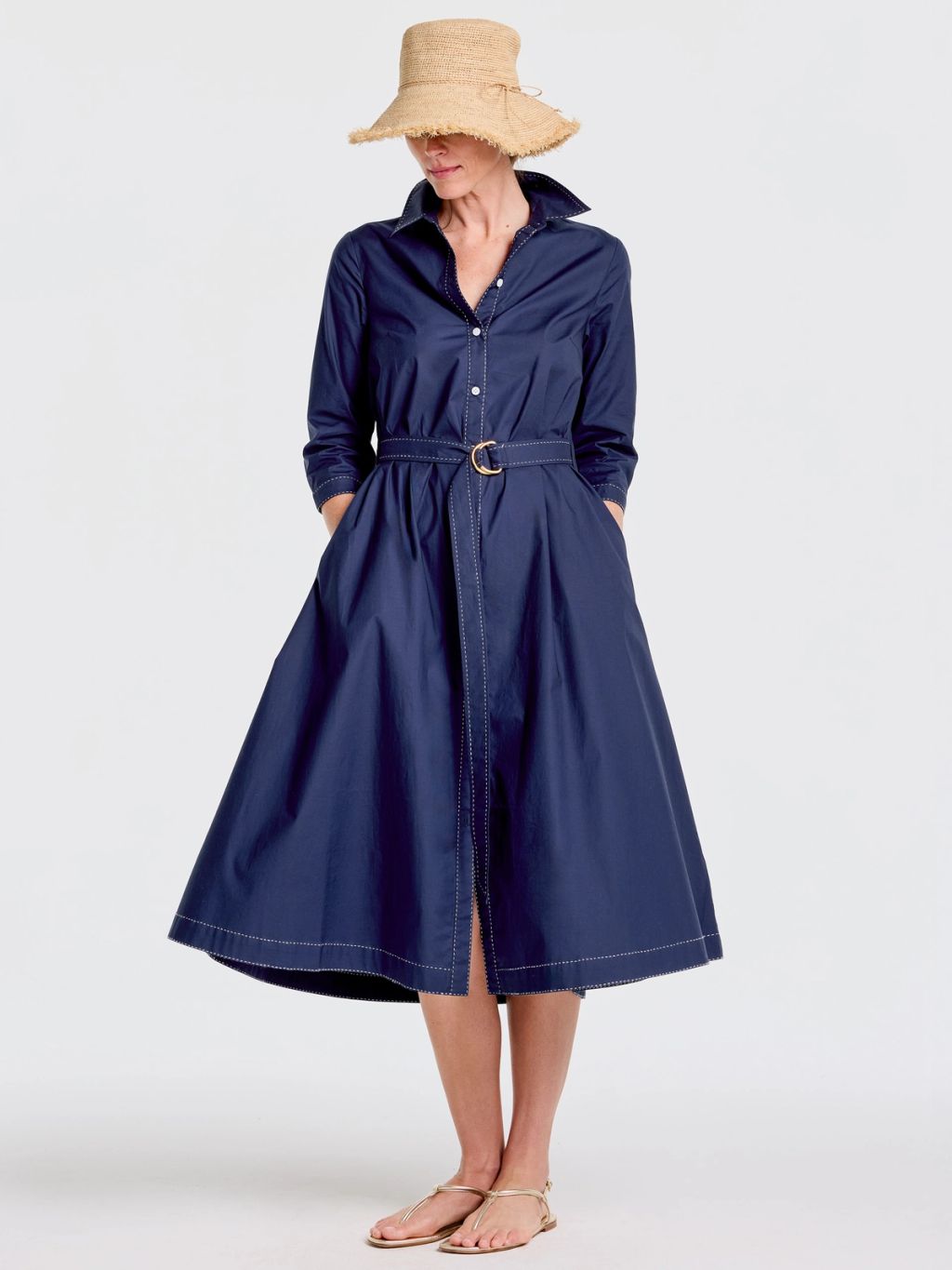 A woman in a navy blue shirtdress with white contrast stitching, a straw hat, and gold sandals stands with hands in her pockets.