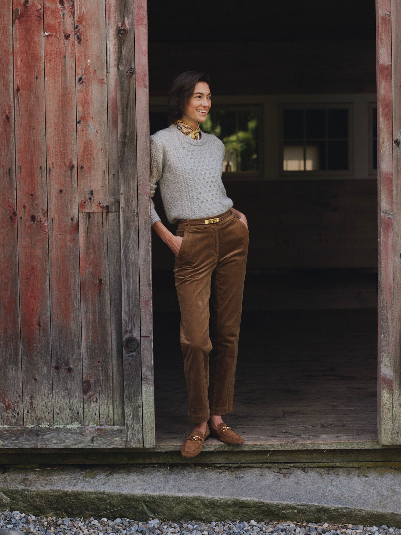 A smiling woman in a grey sweater and brown corduroy pants stands in a rustic barn doorway.