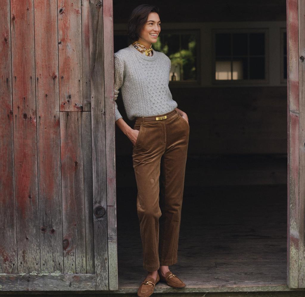 A smiling woman in a grey sweater and brown corduroy pants stands in a rustic barn doorway.