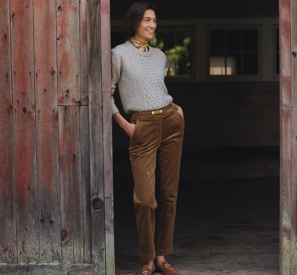 A smiling woman in a grey cable-knit sweater, brown corduroy pants, and a patterned scarf leans in a rustic red doorway.