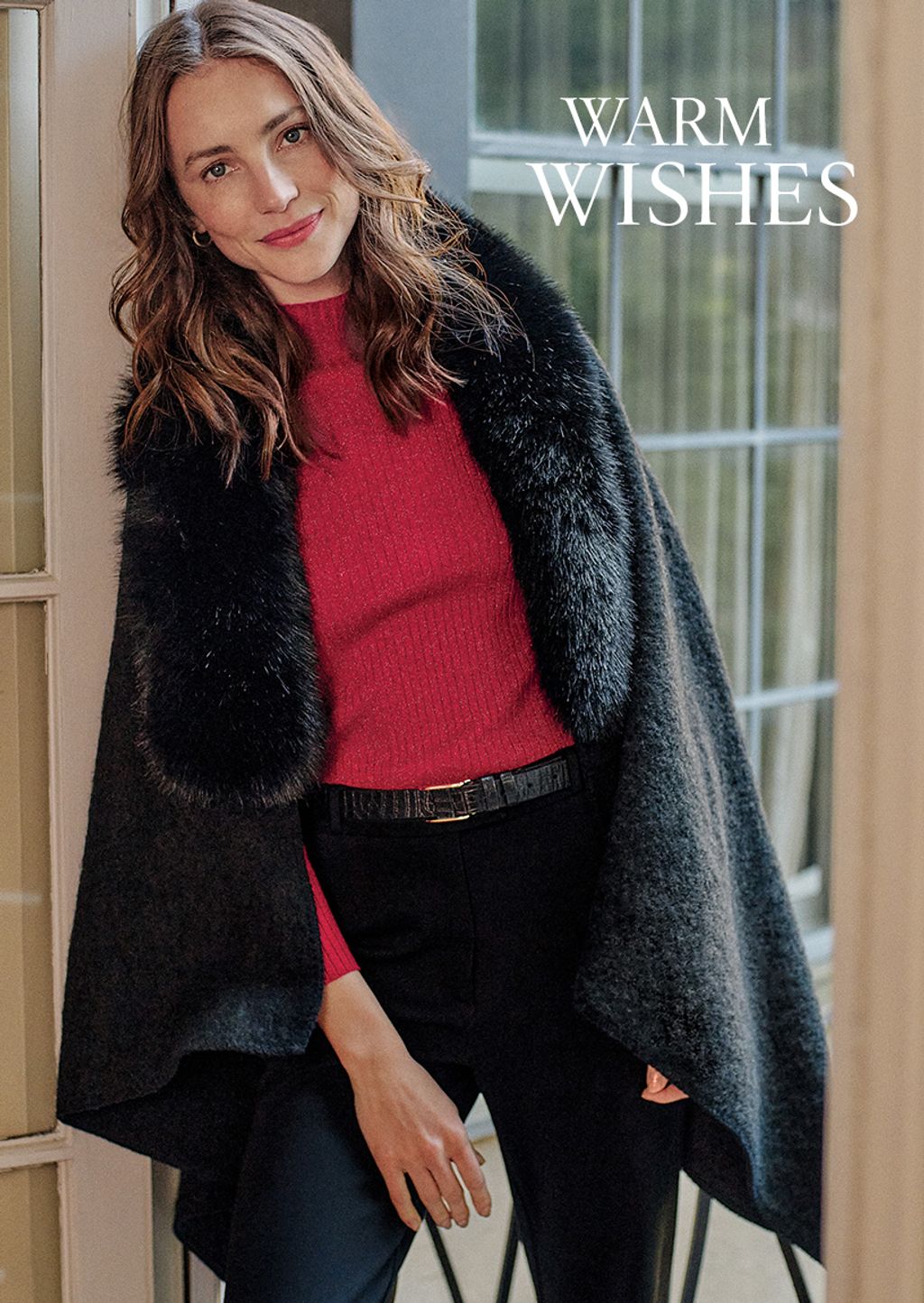 Woman wearing a black cape with faux fur collar, red metallic sweater, a black leather belt and black pants.