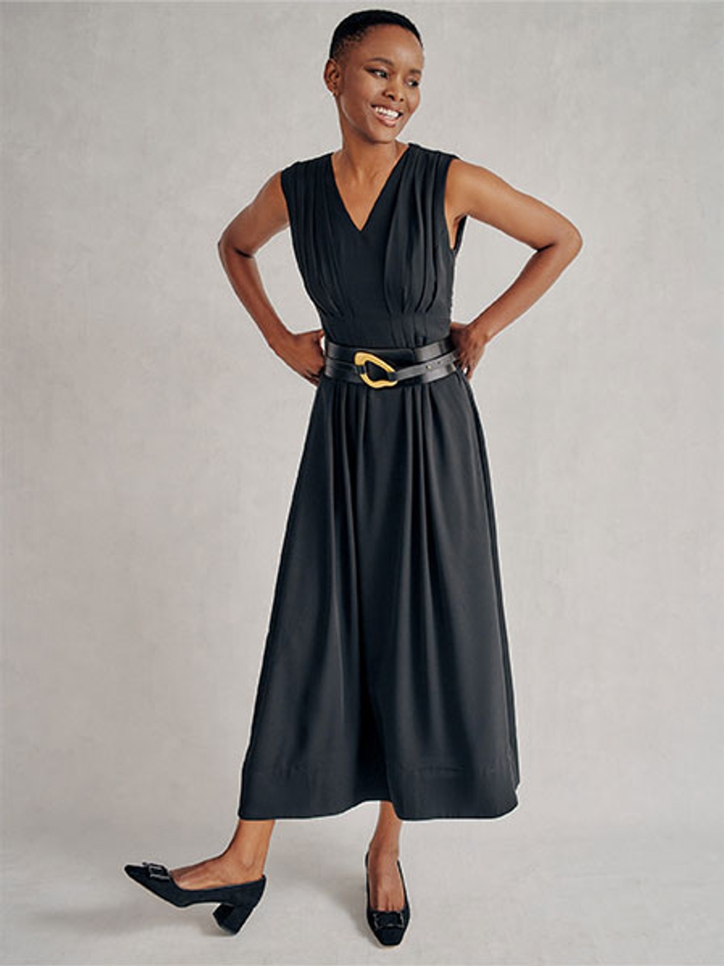 Women standing with hands on her hips wearing a black dress, black heels, and a black leather belt.