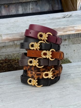 A stack of six belts in various colors and textures, including burgundy, black, brown, and leopard print, all with gold buckles, on a wooden surface.