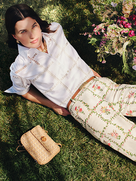 A woman in a white eyelet shirt and floral pants lies on green grass, next to a bouquet of flowers and a woven bag.