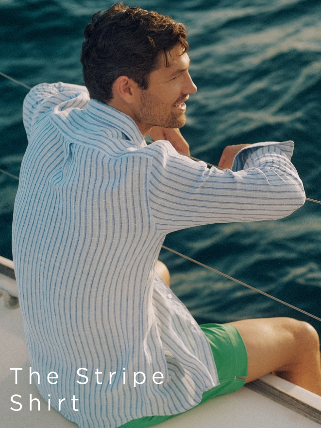 man sitting on edge of boat, sailing on water, wearing blue stripe linen shirt and green swim trunks