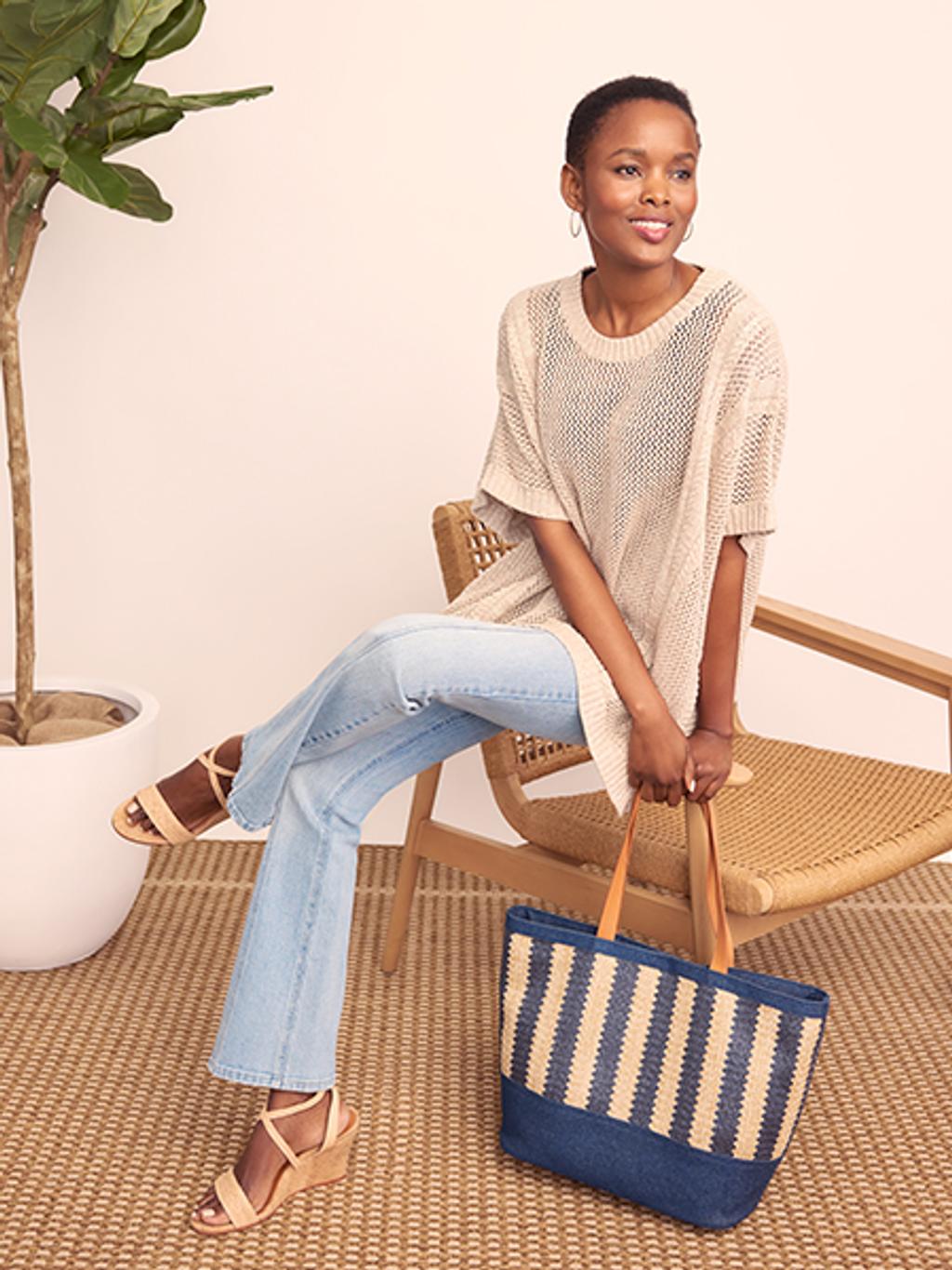 Woman sitting and wearing a knit short sleeve beige top, light wash ankle jeans, leather wedge heels, and carrying a navy striped beach bag.