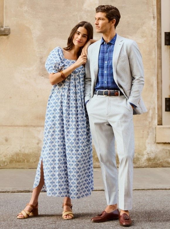 woman and man standing outside against stone building. She is wearing long flowy blouson sleeve white and blue geo pattern dress. He is wearing stone chino slacks with seersucker sport coat over blue tonal check shirt.