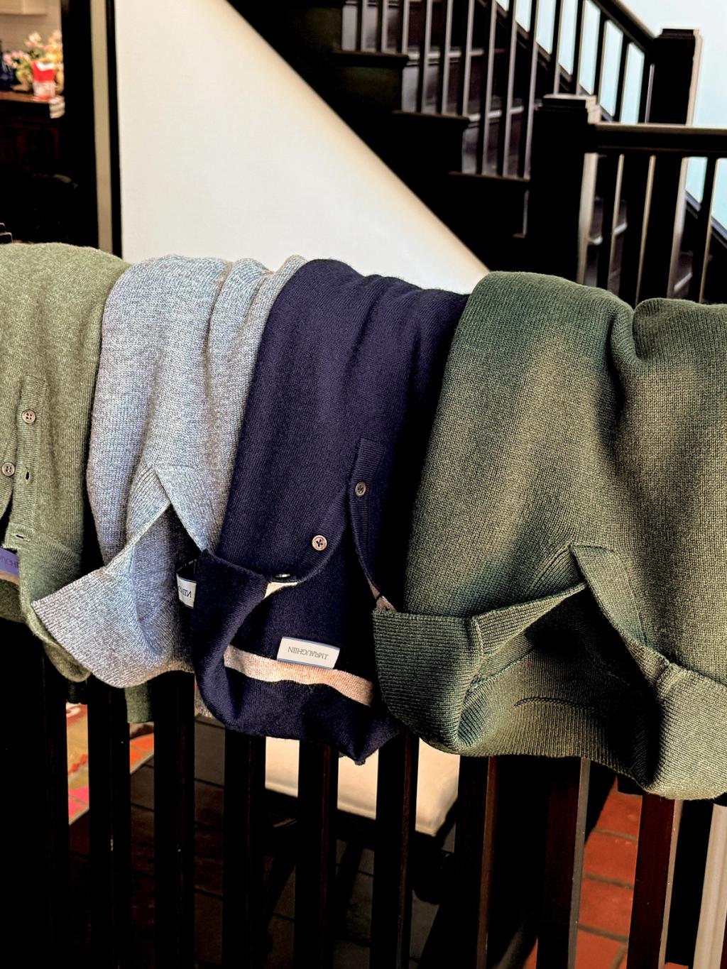 Four cardigans in green, grey, and navy draped over a wooden railing.