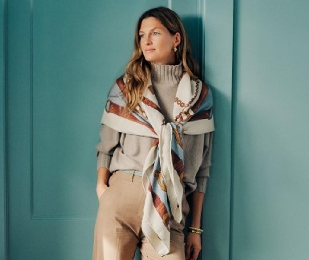 A woman in a beige sweater, tan pants, and a patterned silk scarf leans against a teal wall.