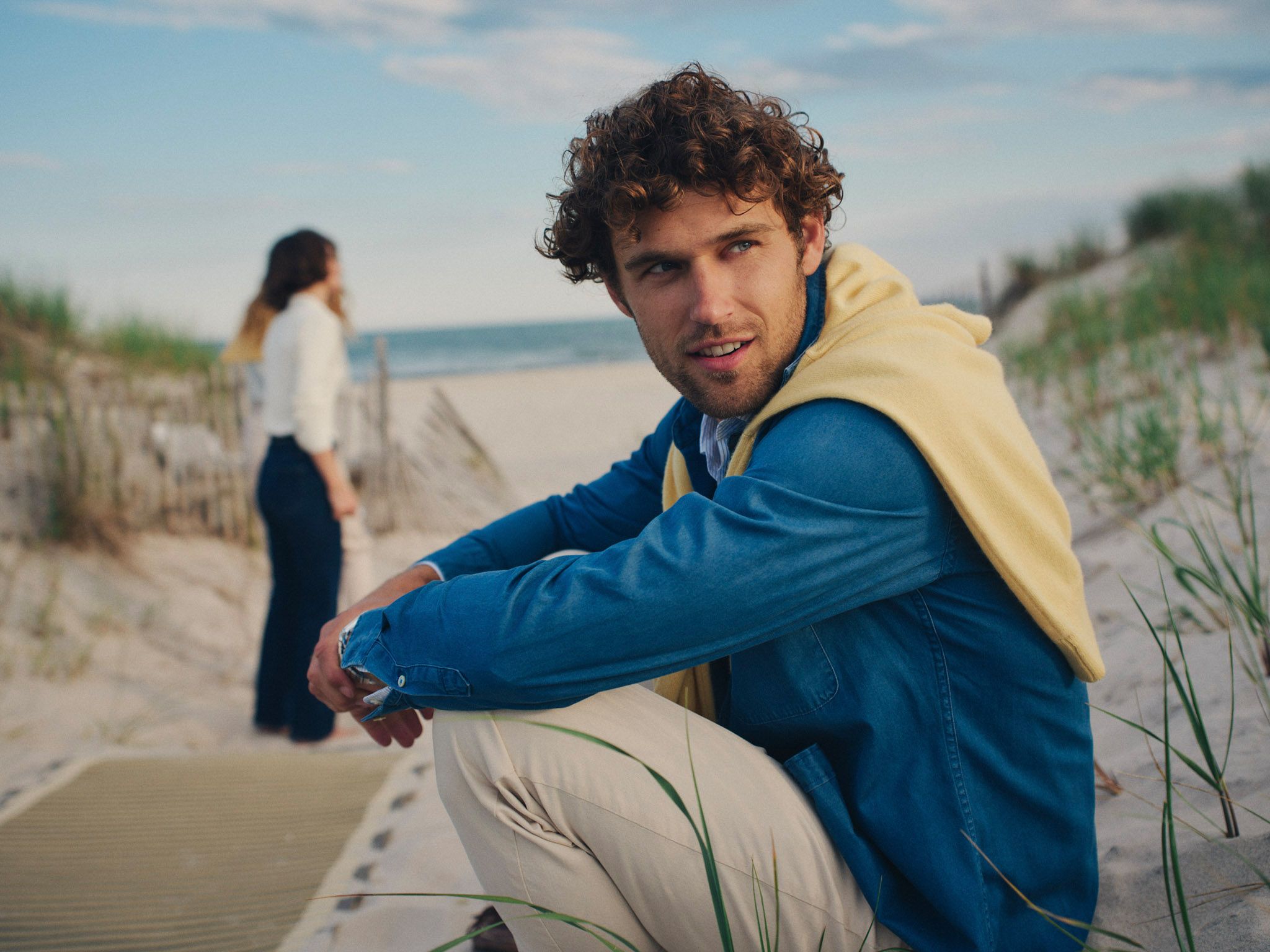A man with curly hair, wearing a blue shirt and yellow sweater, crouches on a sandy beach, looking at the camera, with another person in the background.
