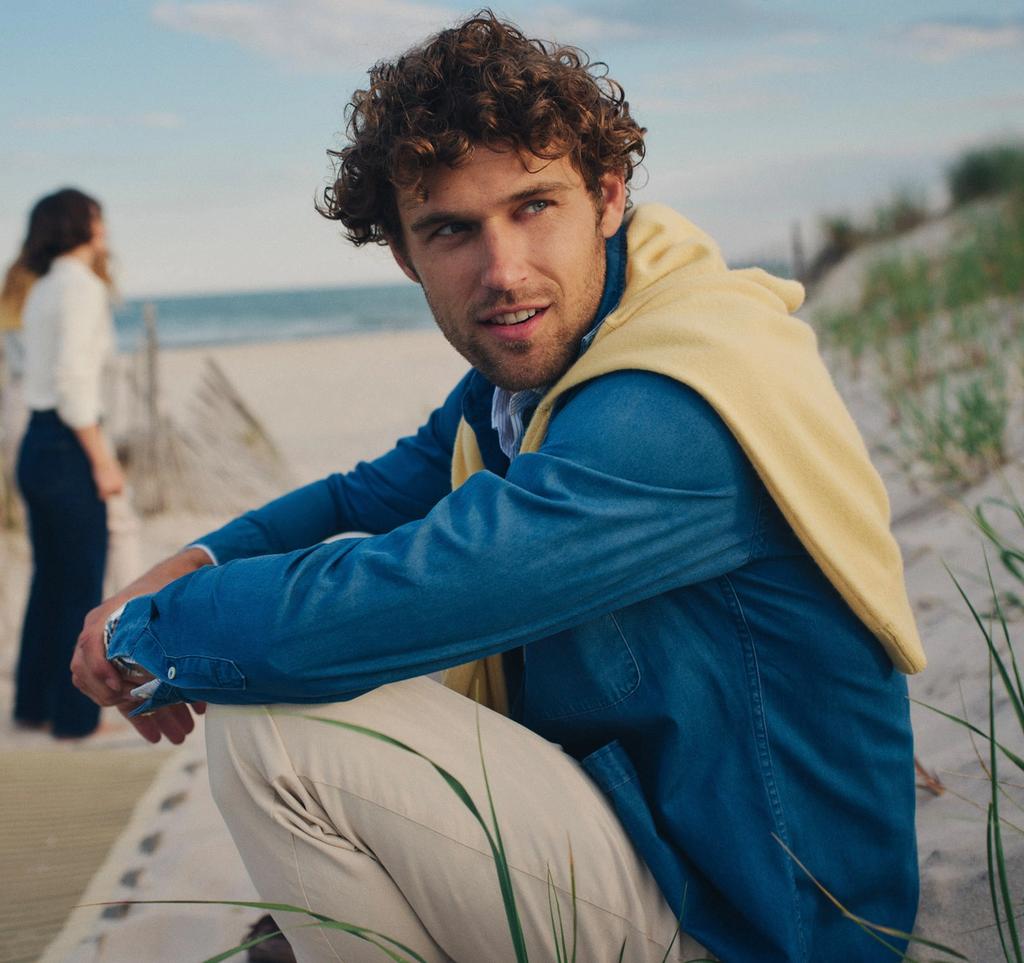 A man with curly hair, wearing a blue shirt and yellow sweater, crouches on a sandy beach, looking at the camera. A woman walks away towards the ocean in the background.