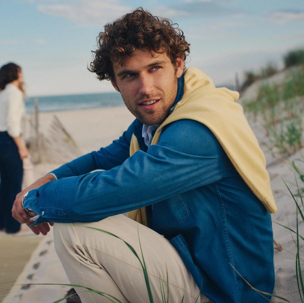 A man with curly hair, wearing a blue shirt and yellow sweater, crouches on a sandy beach, looking at the camera, with another person in the background.