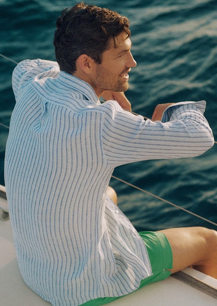 Man in striped shirt and green shorts smiling on a boat, looking at the water.