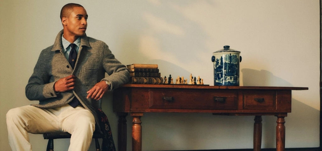 A man in a grey jacket, blue shirt, and tie sits on a stool beside a wooden table with a chessboard and books.