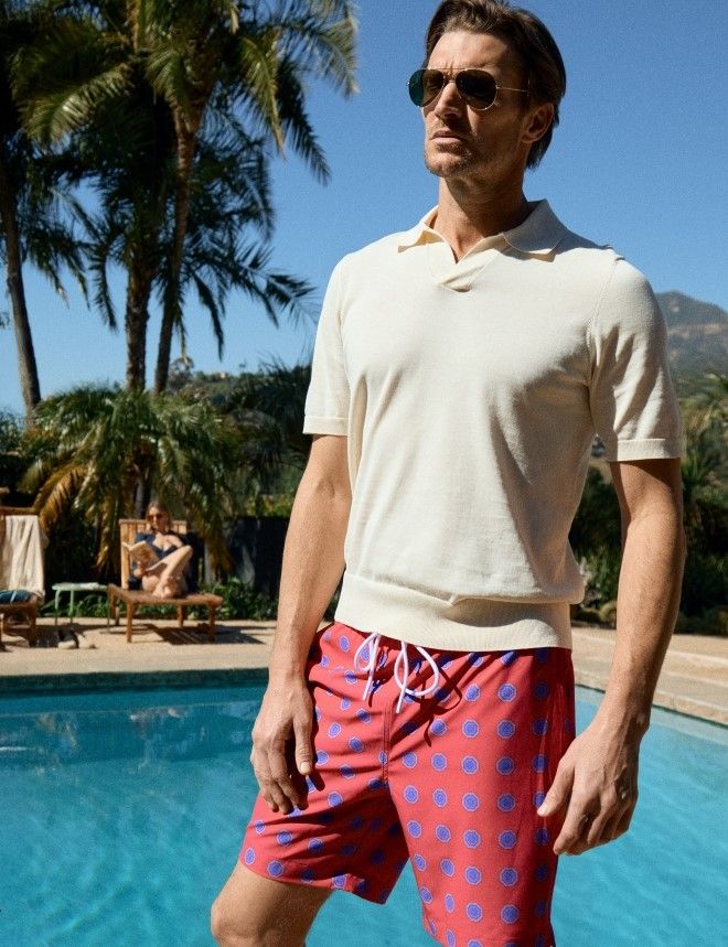 man standing by a pool wearing red swim trunks and yellow knit polo