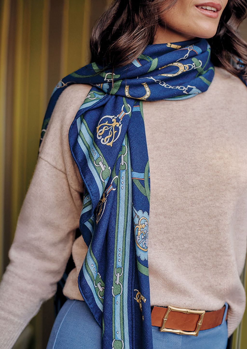 Close up shot of a woman wearing a blue heraldic print scarf, beige sweater, blue pants, and a brown leather belt.