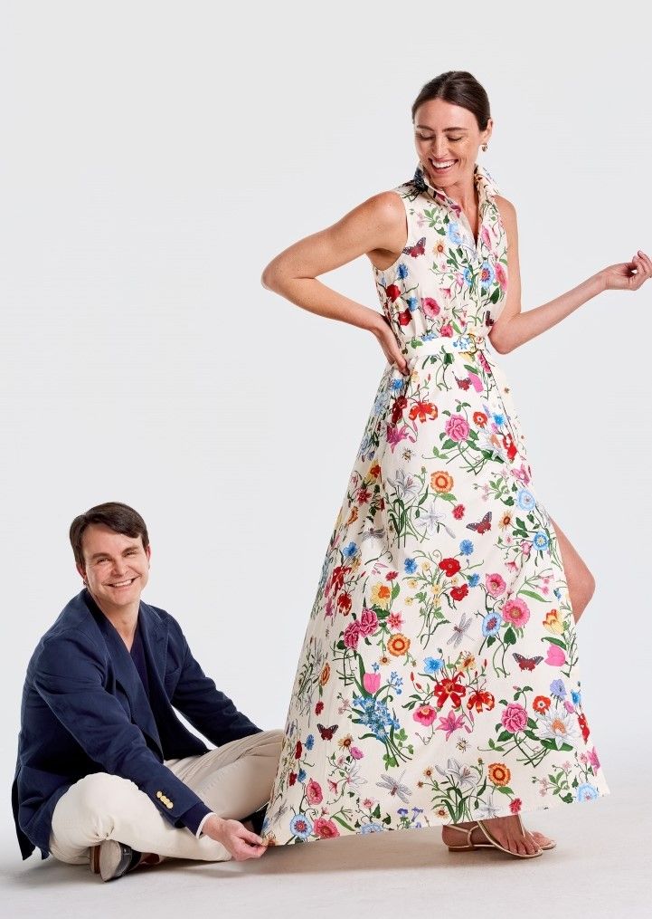 Smiling woman in a floral maxi dress stands while a smiling man sits, holding its hem.