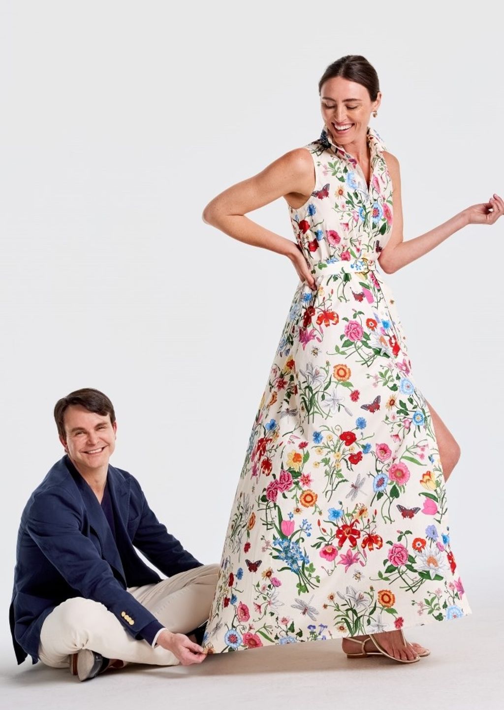 Smiling woman in a floral maxi dress stands while a smiling man sits, holding its hem.