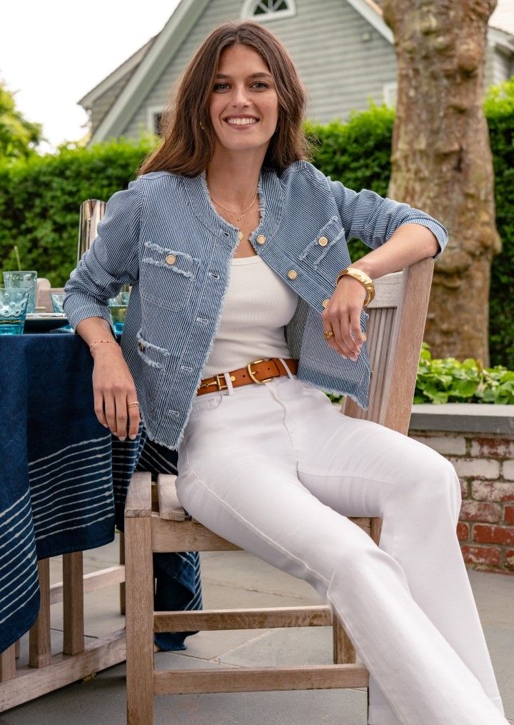 Woman seated at an outside table wearing white jeans, a white tank top and a chambray hairline stripe structured denim jacket