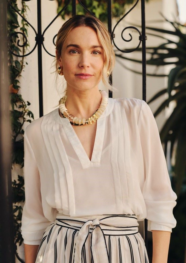 A blonde woman in a sheer white blouse, striped skirt, and a chunky beaded necklace smiles faintly at the camera while standing by a black ornate gate.