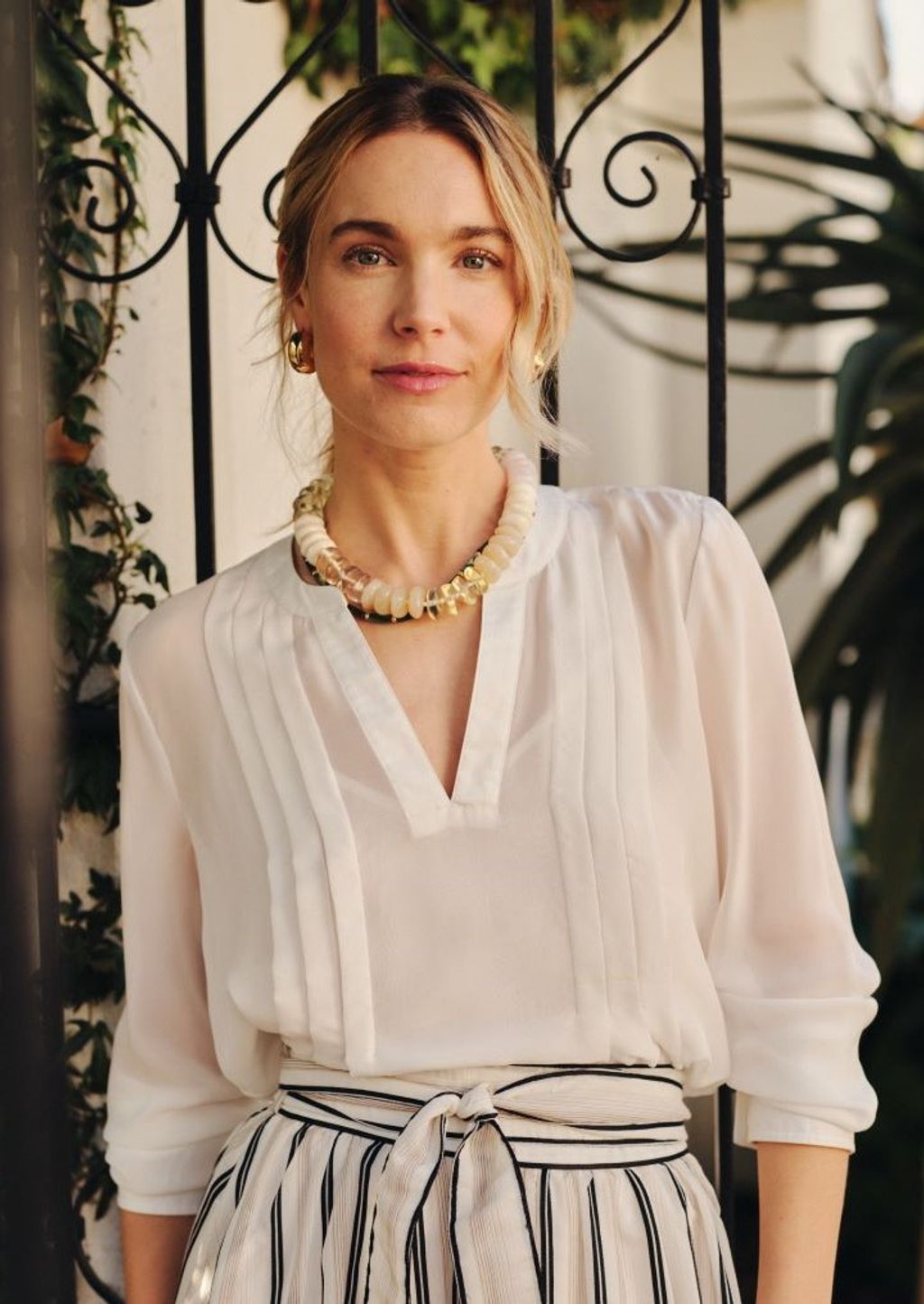 A blonde woman in a sheer white blouse, striped skirt, and a chunky beaded necklace smiles faintly at the camera while standing by a black ornate gate.