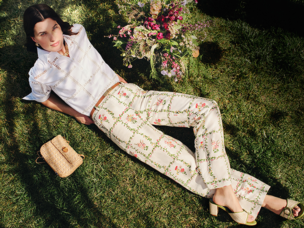 A woman in a white eyelet shirt and floral pants lies on green grass, next to a bouquet of flowers and a woven bag.