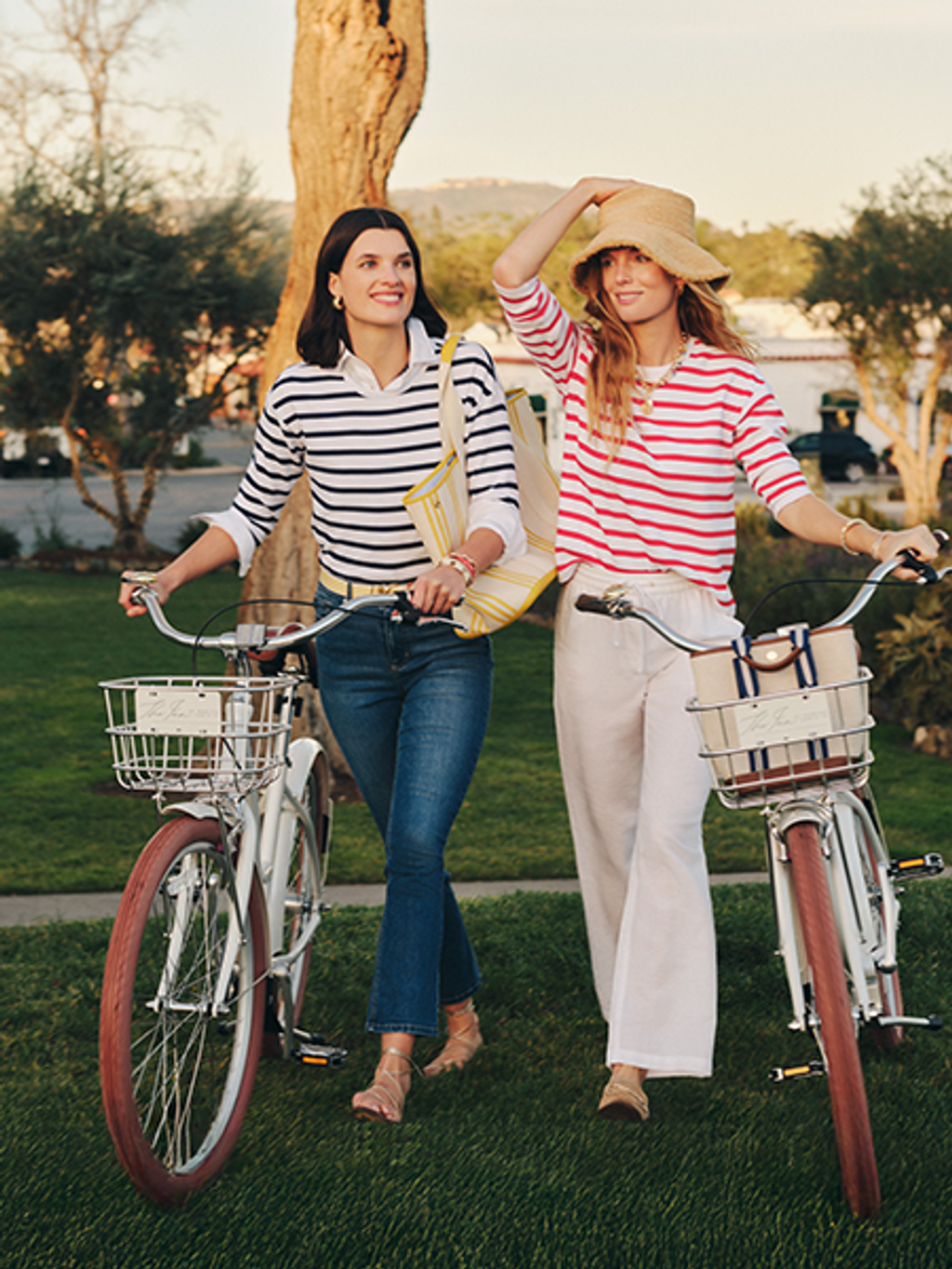 Two women in striped shirts walk their bicycles on a grassy lawn.