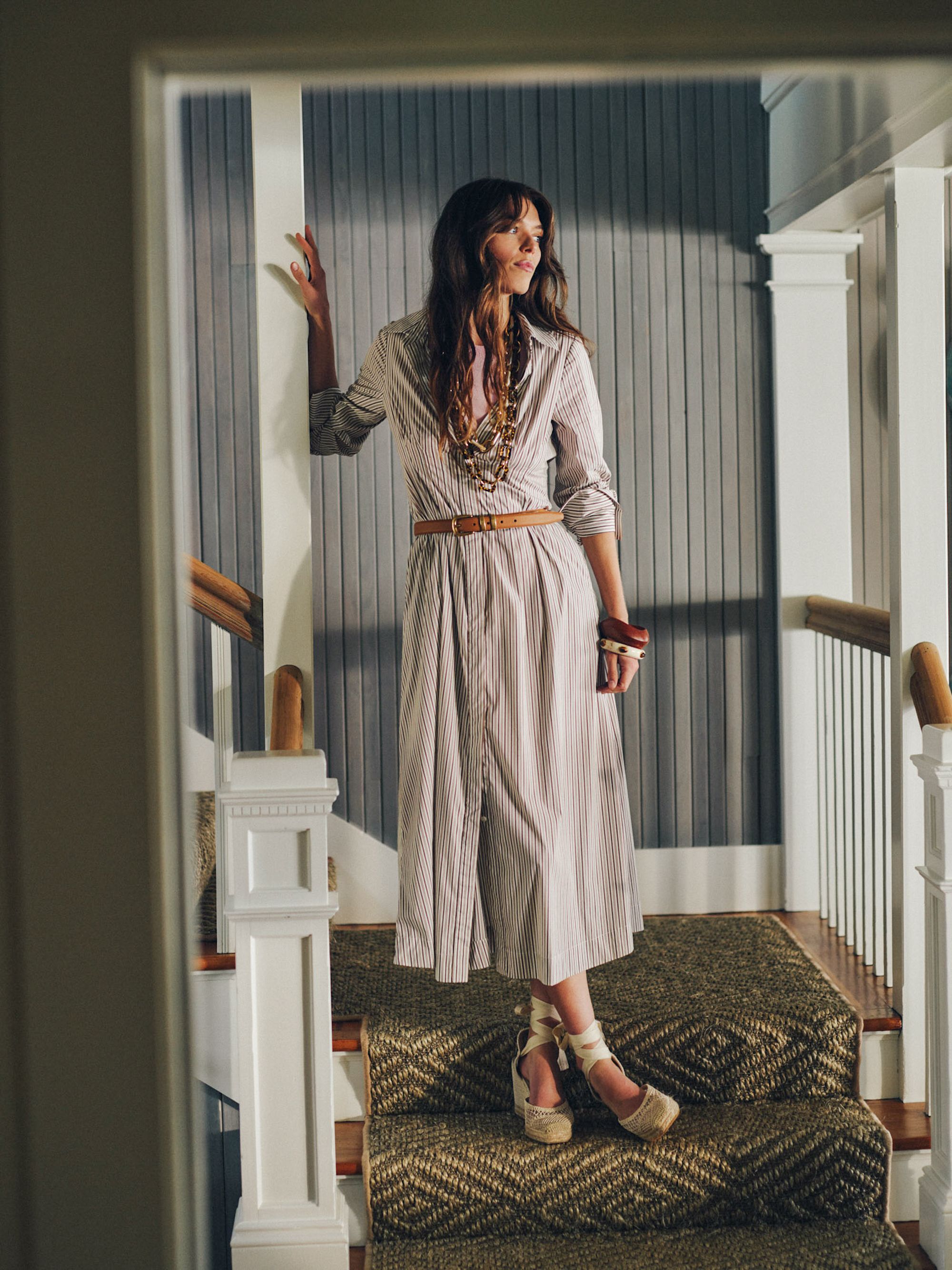 A woman in a striped shirt dress, brown belt, layered necklaces, and espadrille wedges stands on a carpeted staircase.