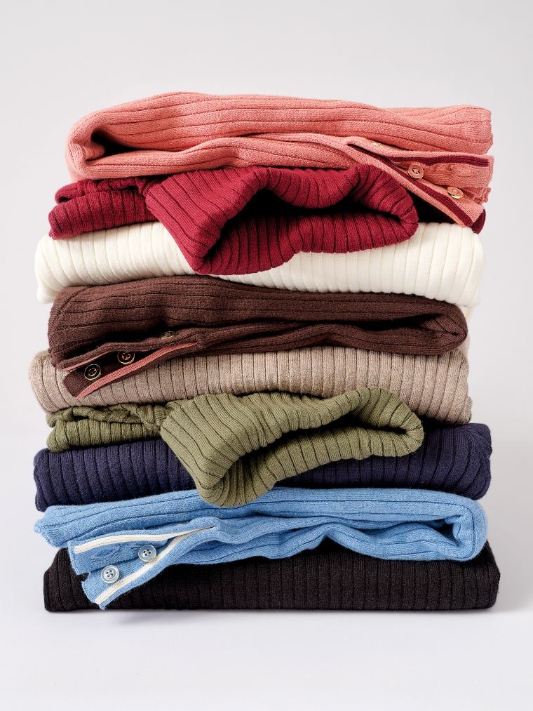 A stack of nine folded ribbed knit sweaters in various colors.