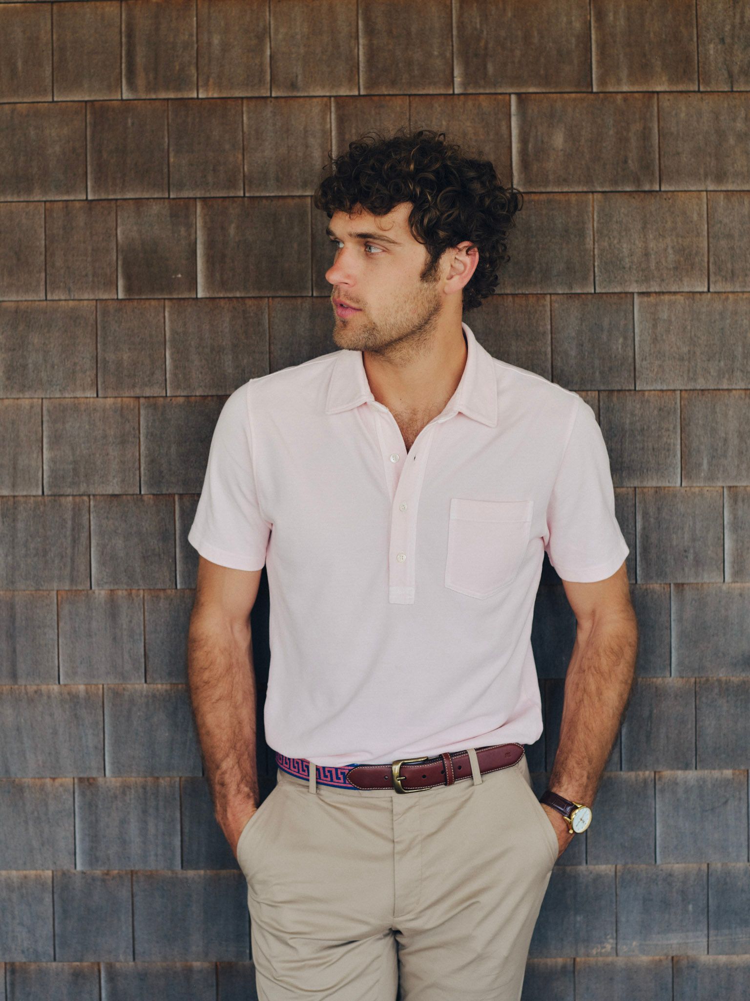 A man in a light pink polo shirt and tan khakis stands against a wooden shingle wall, looking left.