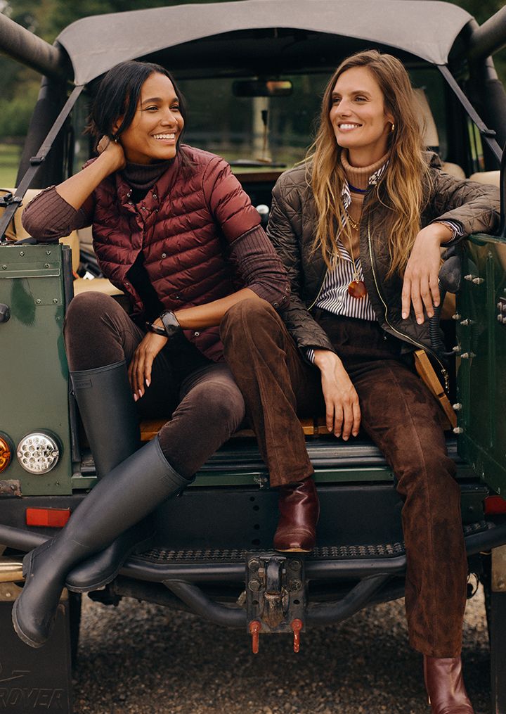 two women sitting in back of trunk, one wearing burgundy puffer jacket, the other a quilted olive jacket with suede pants